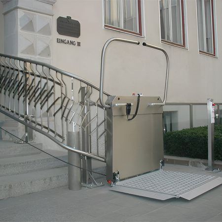 Rollstuhl-Plattformlift an einer geschwungenen Treppe vor einem Gebäude mit dem Schild "Eingang"